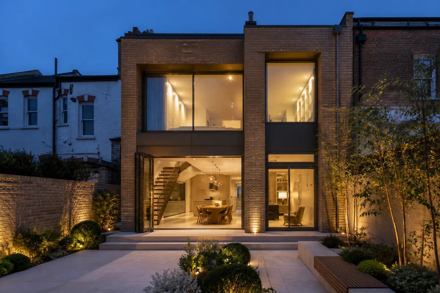 A brick and glass rear extension lit at dusk, opening onto a landscaped garden
